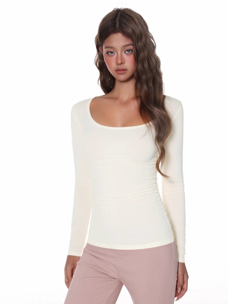 Model wearing a fitted, cream, long-sleeved, scoop neck top with a ribbed texture and ruched side detail.