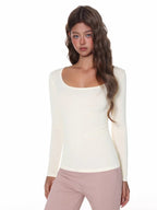 Model wearing a fitted, cream, long-sleeved, scoop neck top with a ribbed texture and ruched side detail.