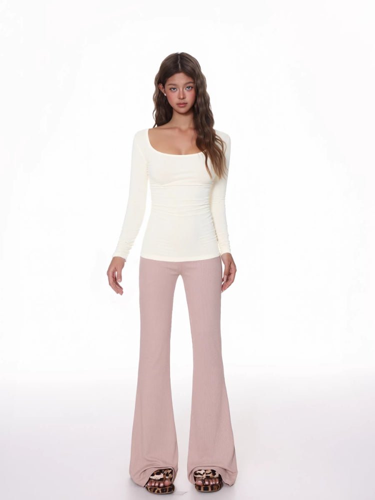 Model wearing a fitted, cream, long-sleeved, scoop neck top with a ribbed texture and ruched side detail, paired with pink flared pants.