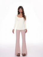 Model wearing a fitted, cream, long-sleeved, scoop neck top with a ribbed texture and ruched side detail, paired with pink flared pants.