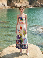 Woman wearing a white sleeveless maxi dress with a colorful graphic print and spaghetti straps, standing on a rock in the ocean