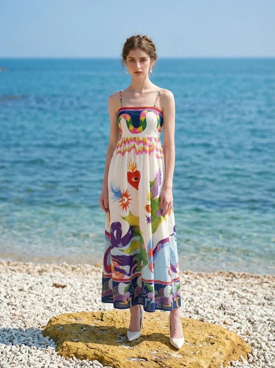 Woman wearing a white sleeveless maxi dress with a colorful graphic print and spaghetti straps, standing on a rock near the ocean
